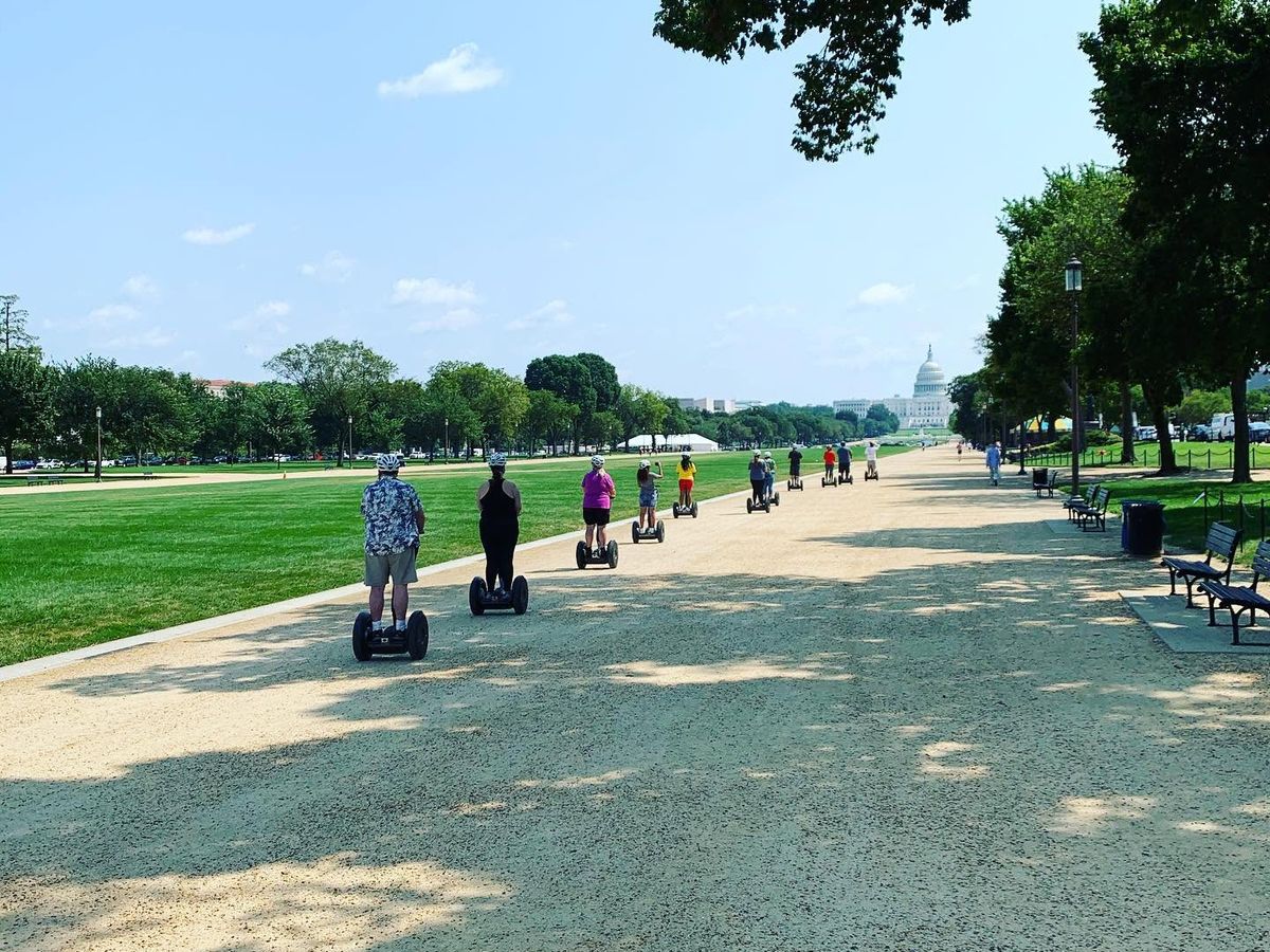 A group of tourists glide along the gravel path of the National Mall on Segways with Capital Segway, heading toward the iconic dome of the US Capitol building under a clear blue summer sky.