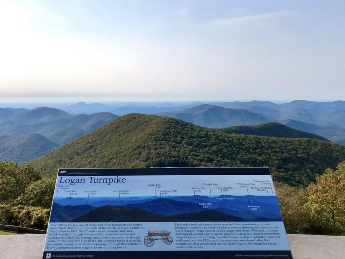 An informational sign about the Logan Turnpike sits in front of a panoramic view of the Blue Ridge Mountains in North Georgia under a clear sky.
