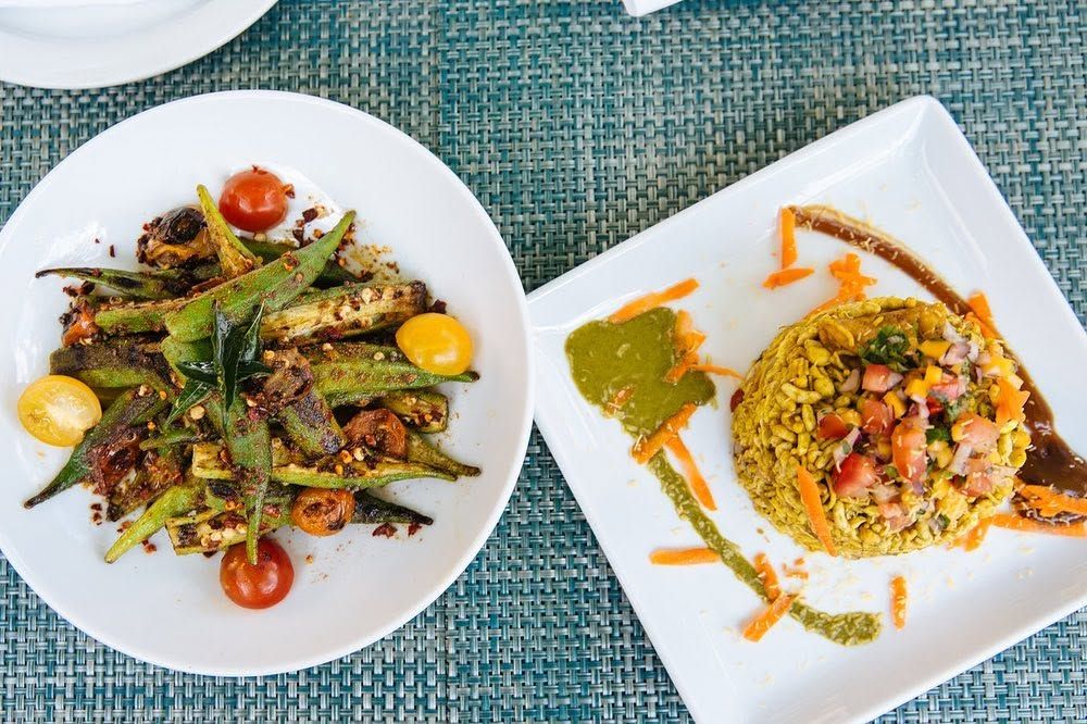 Two vibrant plates rest on a teal woven placemat, one holding blistered okra with cherry tomatoes and curry leaves, the other a mound of puffed rice bhel topped with diced tomato, onion, and shredded carrot beside swirls of green and tamarind chutney. The colorful presentation highlights bold South Indian flavors