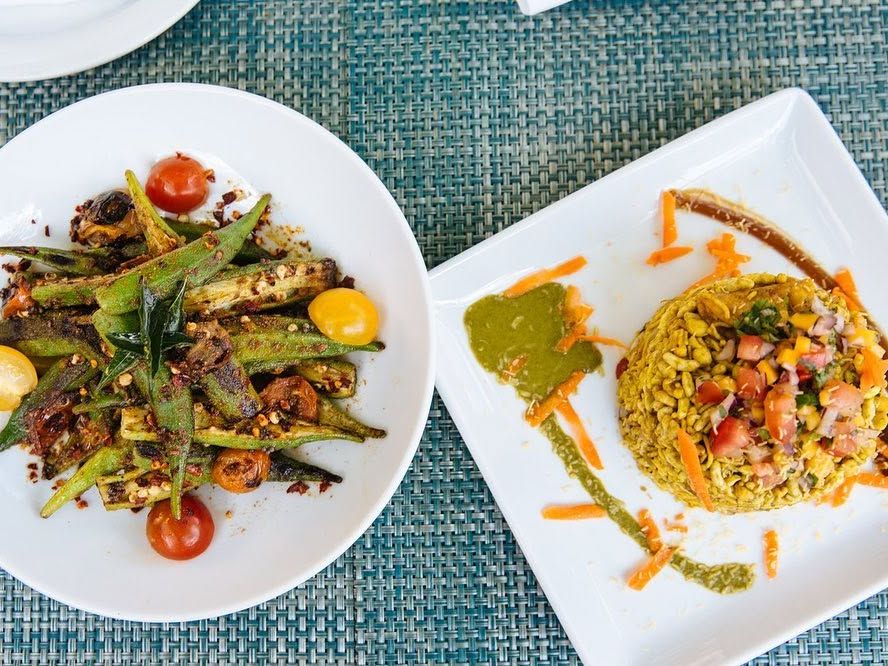 Two vibrant plates rest on a teal woven placemat, one holding blistered okra with cherry tomatoes and curry leaves, the other a mound of puffed rice bhel topped with diced tomato, onion, and shredded carrot beside swirls of green and tamarind chutney. The colorful presentation highlights bold South Indian flavors