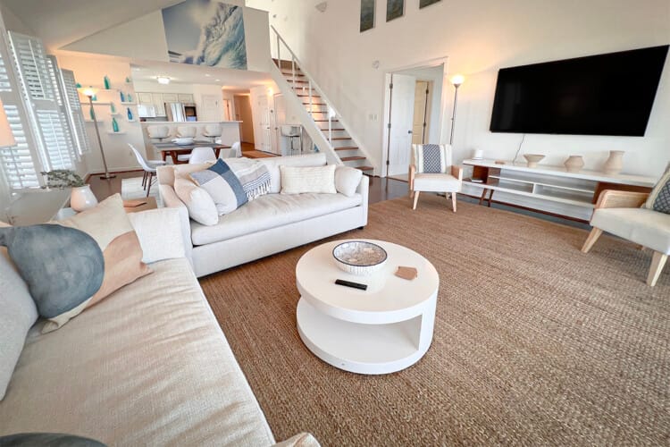 A coastal-inspired living room with cream sofas, woven rug, and ocean artwork above the staircase. The open layout connects to a bright kitchen, creating a breezy, relaxed atmosphere.