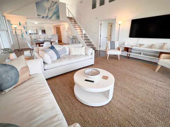 A coastal-inspired living room with cream sofas, woven rug, and ocean artwork above the staircase. The open layout connects to a bright kitchen, creating a breezy, relaxed atmosphere.