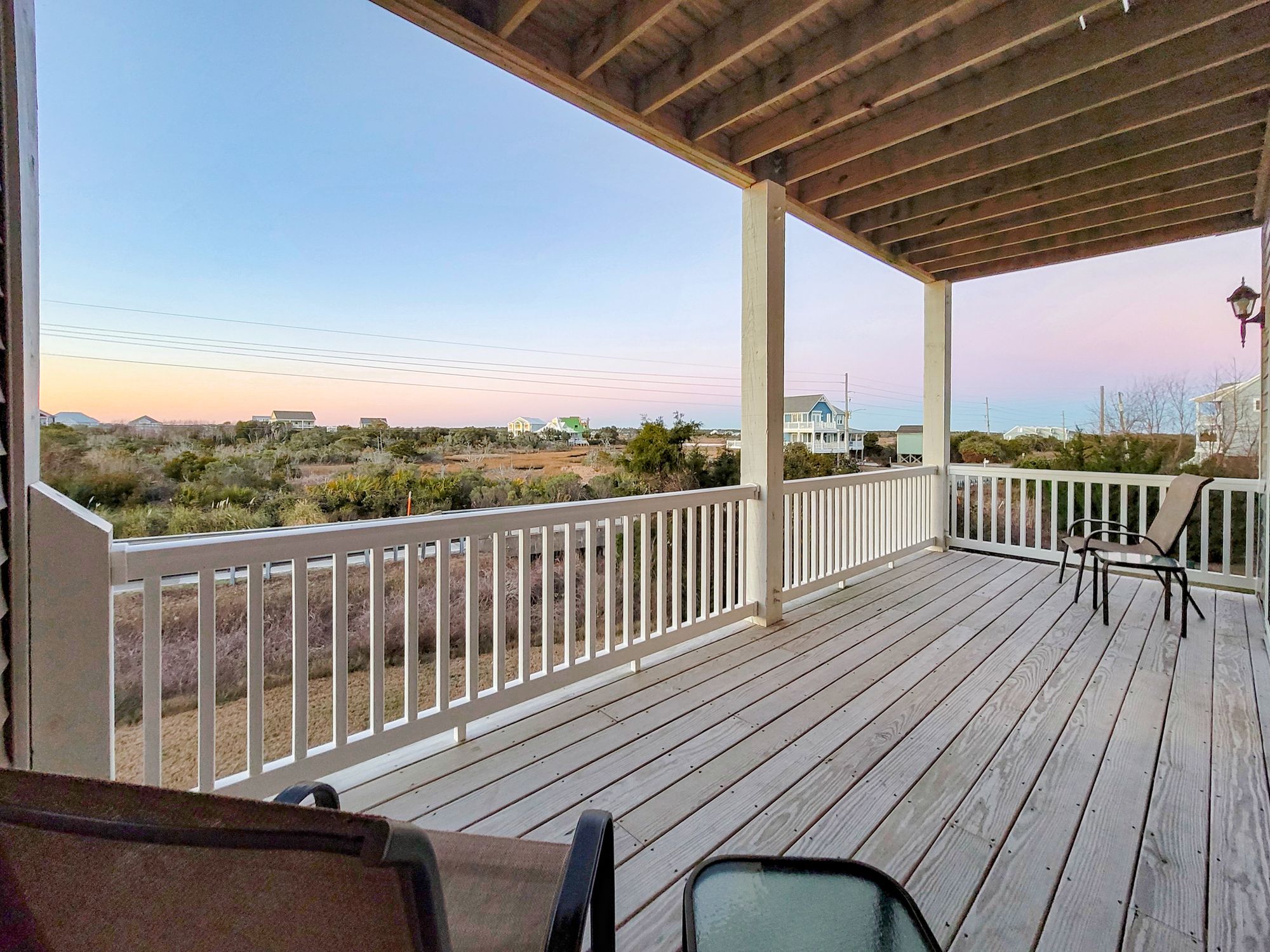 A spacious wooden deck with white railings and outdoor seating, overlooking a coastal landscape with scattered beach houses under a soft pastel sunset sky.