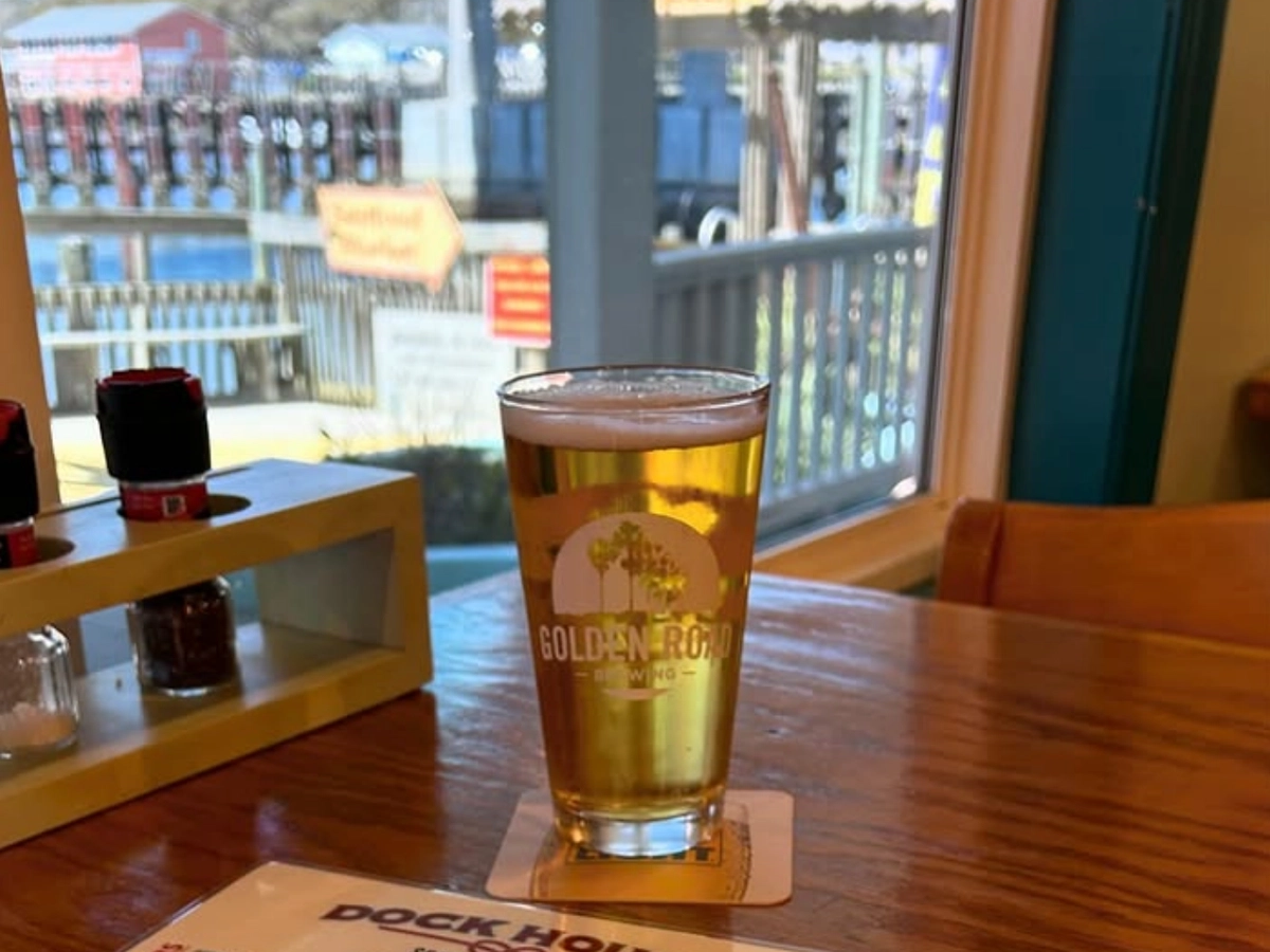A pint of Golden Road beer sits on a wooden table beside a Dock House menu, with a waterfront seafood shack visible through the sunny window.