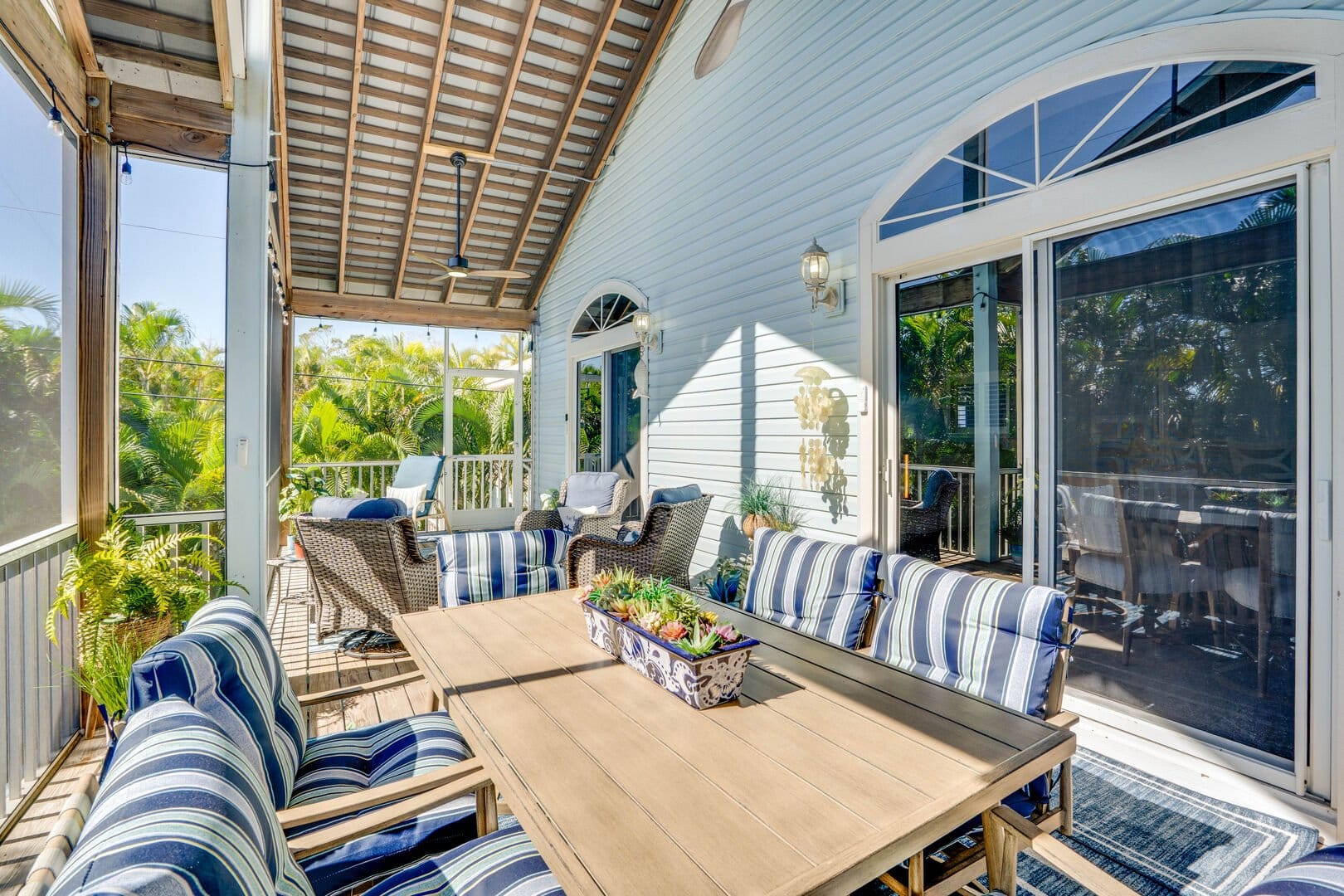 Covered coastal veranda with striped cushioned seating, wooden dining table, and lush tropical greenery surrounding the screened outdoor space.