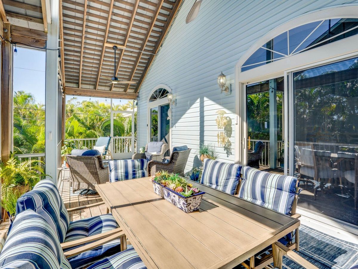 Covered coastal veranda with striped cushioned seating, wooden dining table, and lush tropical greenery surrounding the screened outdoor space.