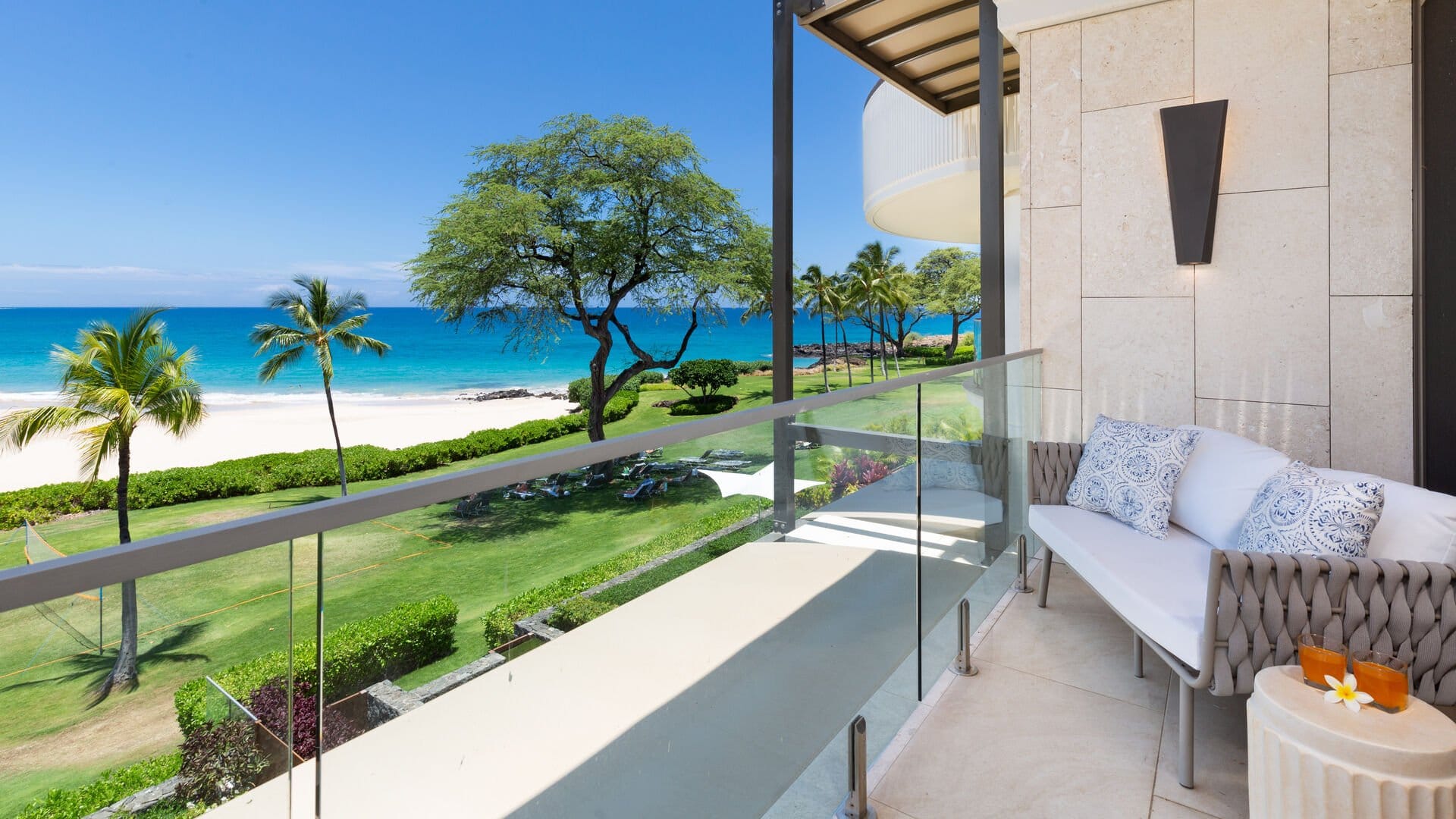 A furnished lanai at Hapuna Sunsets offers a stunning oceanfront view of Hapuna Beach on the Big Island of Hawaii, with turquoise Pacific waters, white sand, and swaying palm trees visible beyond the glass-railed balcony. A plumeria blossom rests on the side table beside a cushioned wicker sofa, setting the scene for relaxed luxury living.