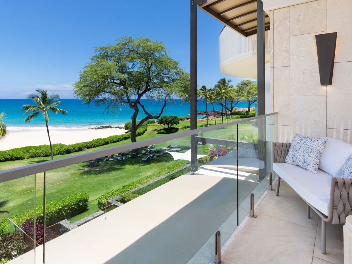 A furnished lanai at Hapuna Sunsets offers a stunning oceanfront view of Hapuna Beach on the Big Island of Hawaii, with turquoise Pacific waters, white sand, and swaying palm trees visible beyond the glass-railed balcony. A plumeria blossom rests on the side table beside a cushioned wicker sofa, setting the scene for relaxed luxury living.