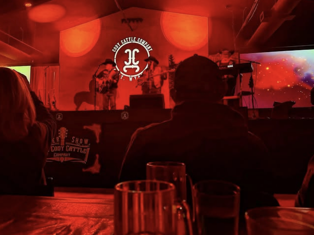 Audience view of a live country music performance at Cody Cattle Company, with a red-lit stage, musicians in cowboy hats, and drinks on the table in the foreground.