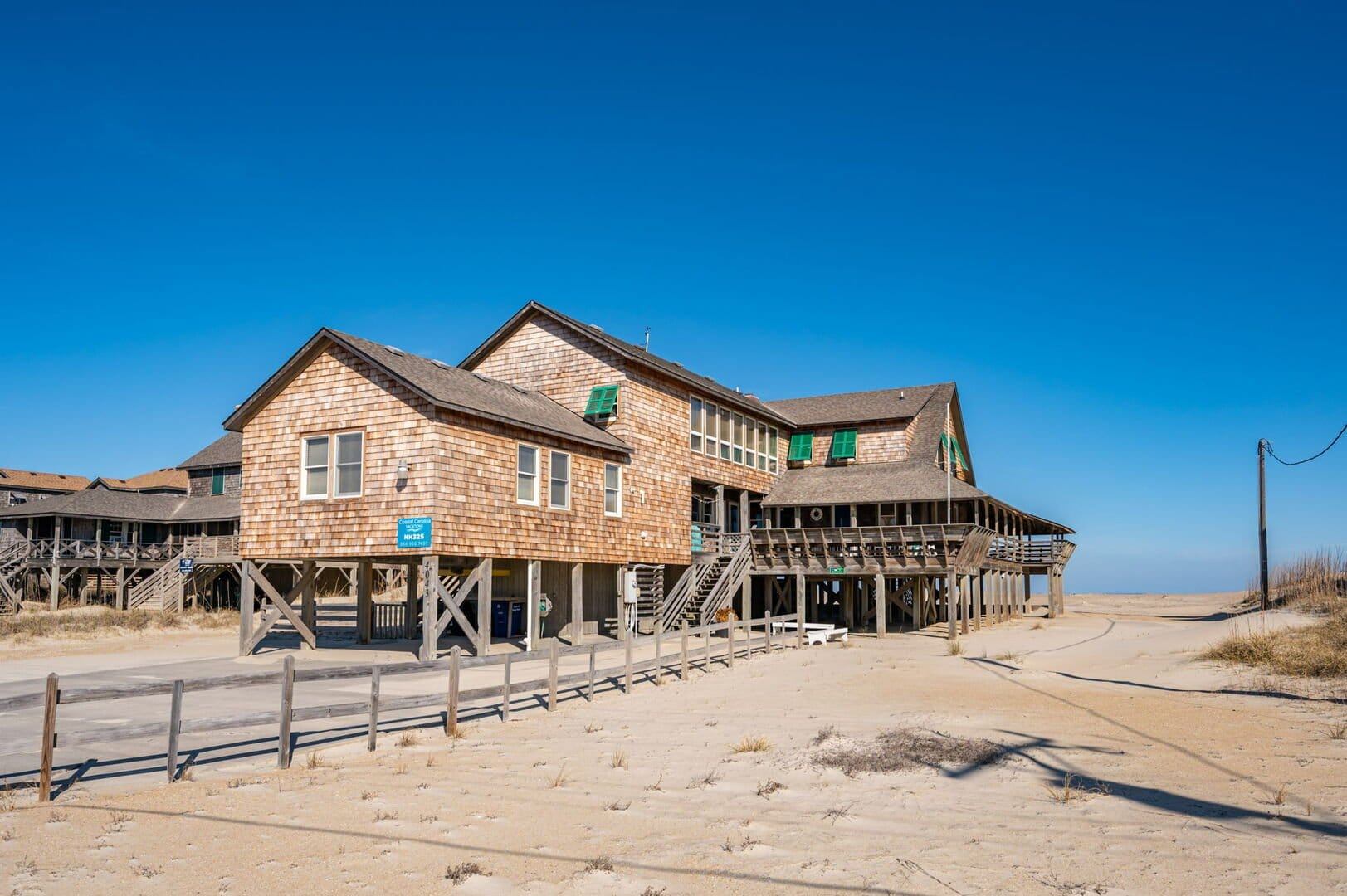 "A rustic beachfront home built on stilts with weathered cedar shake siding and green shutters, situated on a sandy dune under a clear blue sky. The house features multiple interconnected sections, elevated wooden walkways, and wraparound porches with railings. A wooden fence lines the sandy path leading toward the ocean, and utility poles stand in the background. The surrounding area is open, with dunes and sparse vegetation blending into the coastal landscape.
