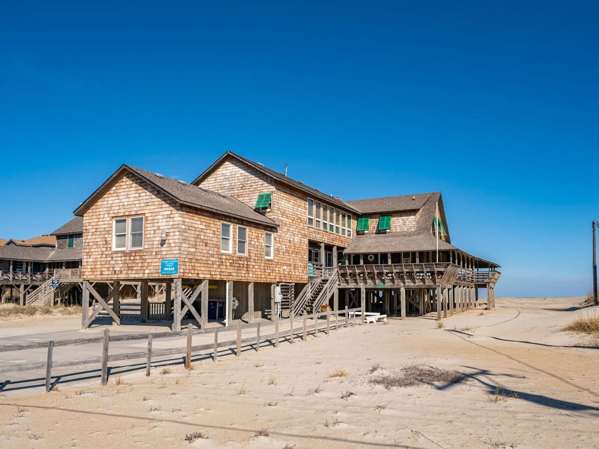 "A rustic beachfront home built on stilts with weathered cedar shake siding and green shutters, situated on a sandy dune under a clear blue sky. The house features multiple interconnected sections, elevated wooden walkways, and wraparound porches with railings. A wooden fence lines the sandy path leading toward the ocean, and utility poles stand in the background. The surrounding area is open, with dunes and sparse vegetation blending into the coastal landscape.
