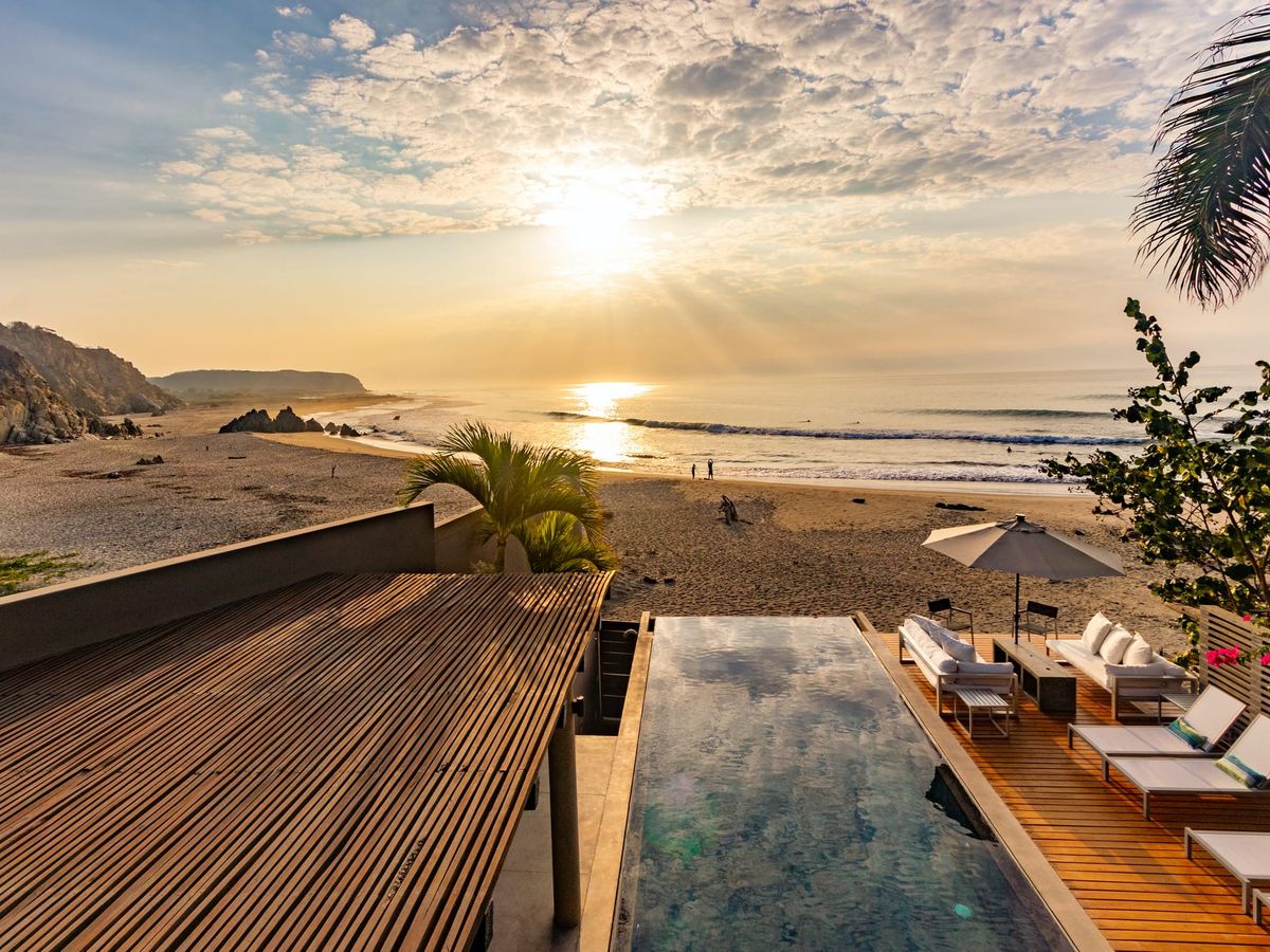 Luxury oceanfront villa with a private infinity pool overlooking a golden sandy beach at sunset. The spacious deck features lounge chairs and a shaded seating area, perfect for relaxing while listening to the waves. This beachfront retreat offers stunning coastal views and a peaceful escape by the sea.