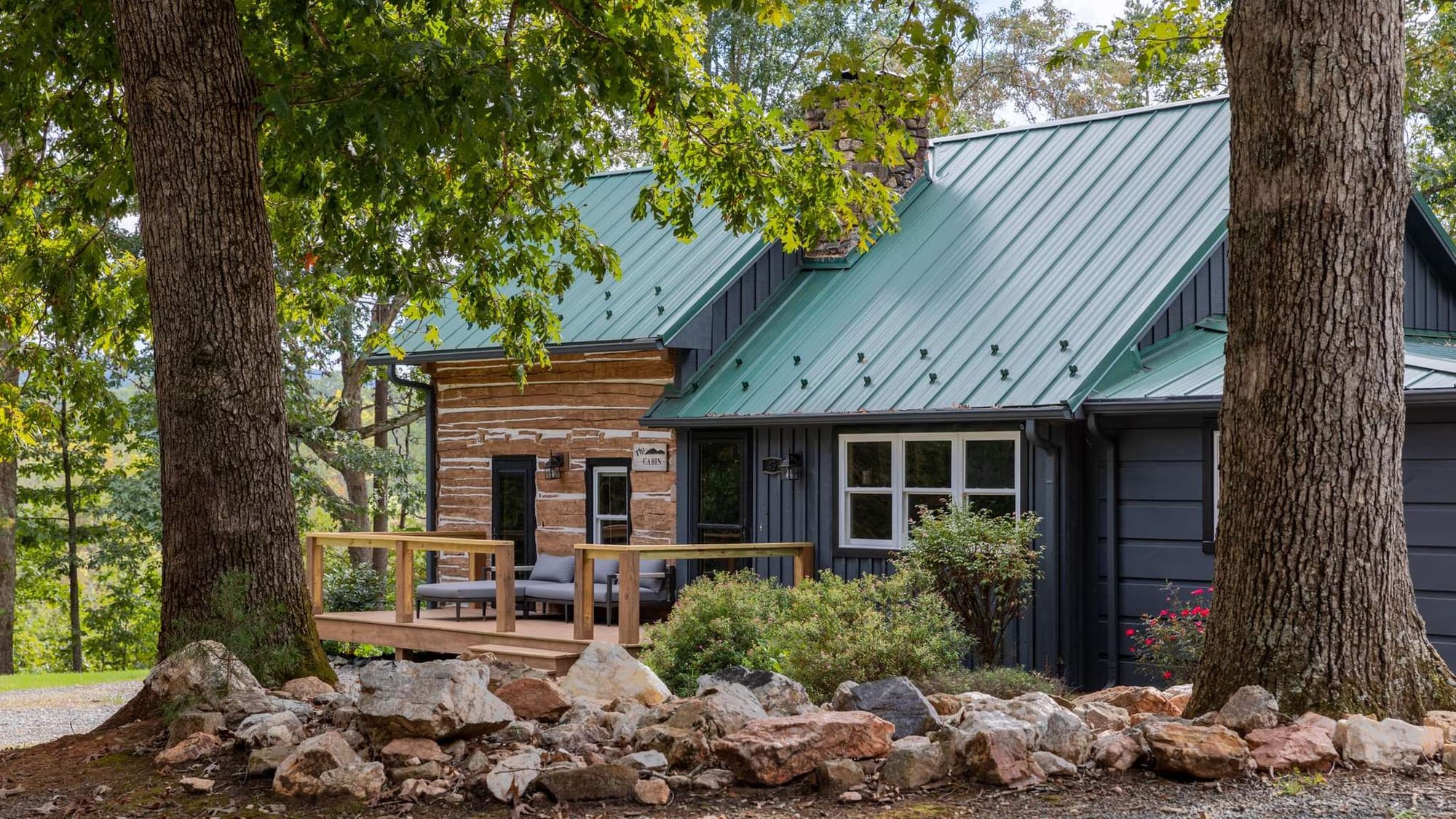 A renovated log cabin with a teal metal roof and charcoal board-and-batten siding sits nestled between two towering oak trees, with a wood-railed porch furnished with outdoor seating. Rocky landscaping and mature foliage frame the cabin's rustic-modern facade.