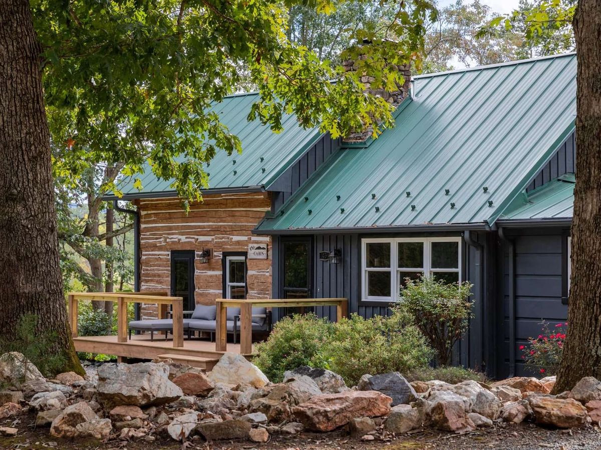 A renovated log cabin with a teal metal roof and charcoal board-and-batten siding sits nestled between two towering oak trees, with a wood-railed porch furnished with outdoor seating. Rocky landscaping and mature foliage frame the cabin's rustic-modern facade.