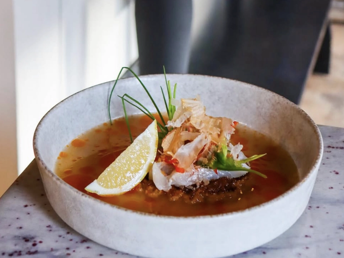 A bowl of flavorful broth topped with a lemon wedge, shredded bonito flakes, herbs, chili oil, and what appears to be a savory protein or patty at the center, served in a minimalist ceramic bowl.