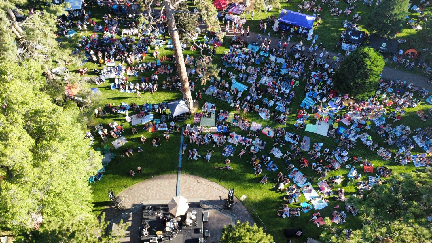 Aerial view of a large outdoor crowd enjoying live music in a park surrounded by tall trees. Blankets, food stalls, and colorful tents create a fun, laid-back summer evening atmosphere at Bend’s Munch & Music event.
