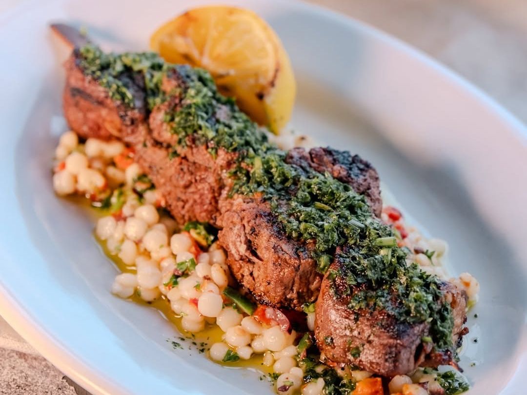 Juicy grilled beef skewers with chimichurri sauce and pearl couscous, plated beside a grilled lemon. A bold and flavorful dish served in a stylish coastal setting.
