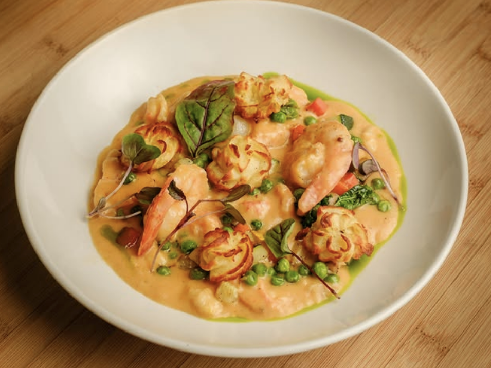 A creamy shrimp and vegetable medley served in a rich orange sauce, topped with piped and roasted duchess potatoes and garnished with microgreens, presented in a white bowl on a wooden surface.