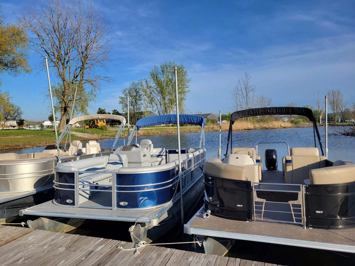 Two pontoon boats with canopy tops are docked side by side at a wooden pier on a calm lake under a bright blue sky. Budding spring trees and a grassy shoreline line the water in the background on a clear, sunny day. 