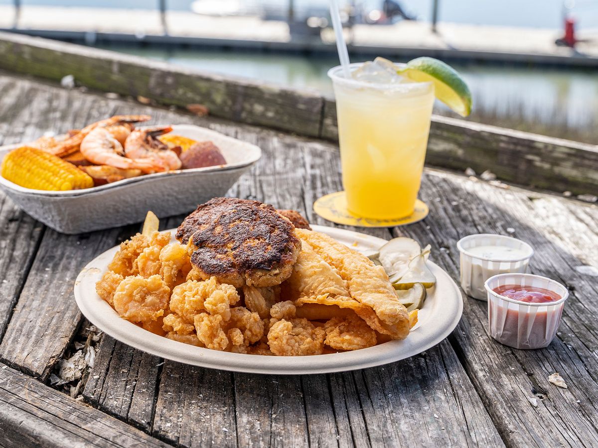 A seafood platter with fried shrimp, hushpuppies, and corn sits on a wooden table near the water at Bowens Island. A cold drink with lime adds to the classic Lowcountry meal. This waterfront spot is known for fresh seafood and marsh views.
