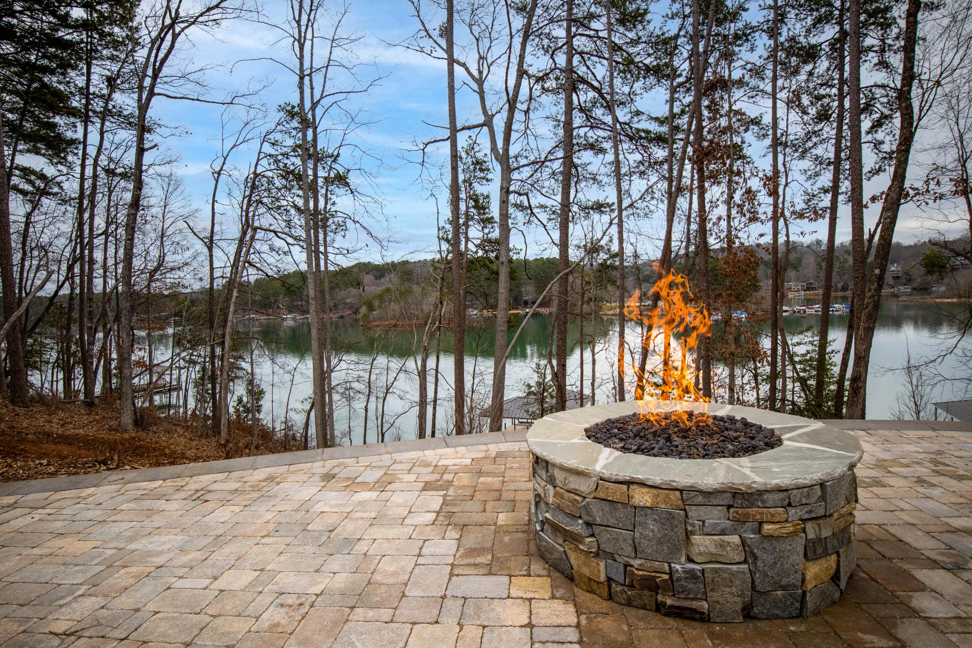 Stone fire pit with flames burning on a paved patio overlooking a scenic lake surrounded by trees and distant houses.