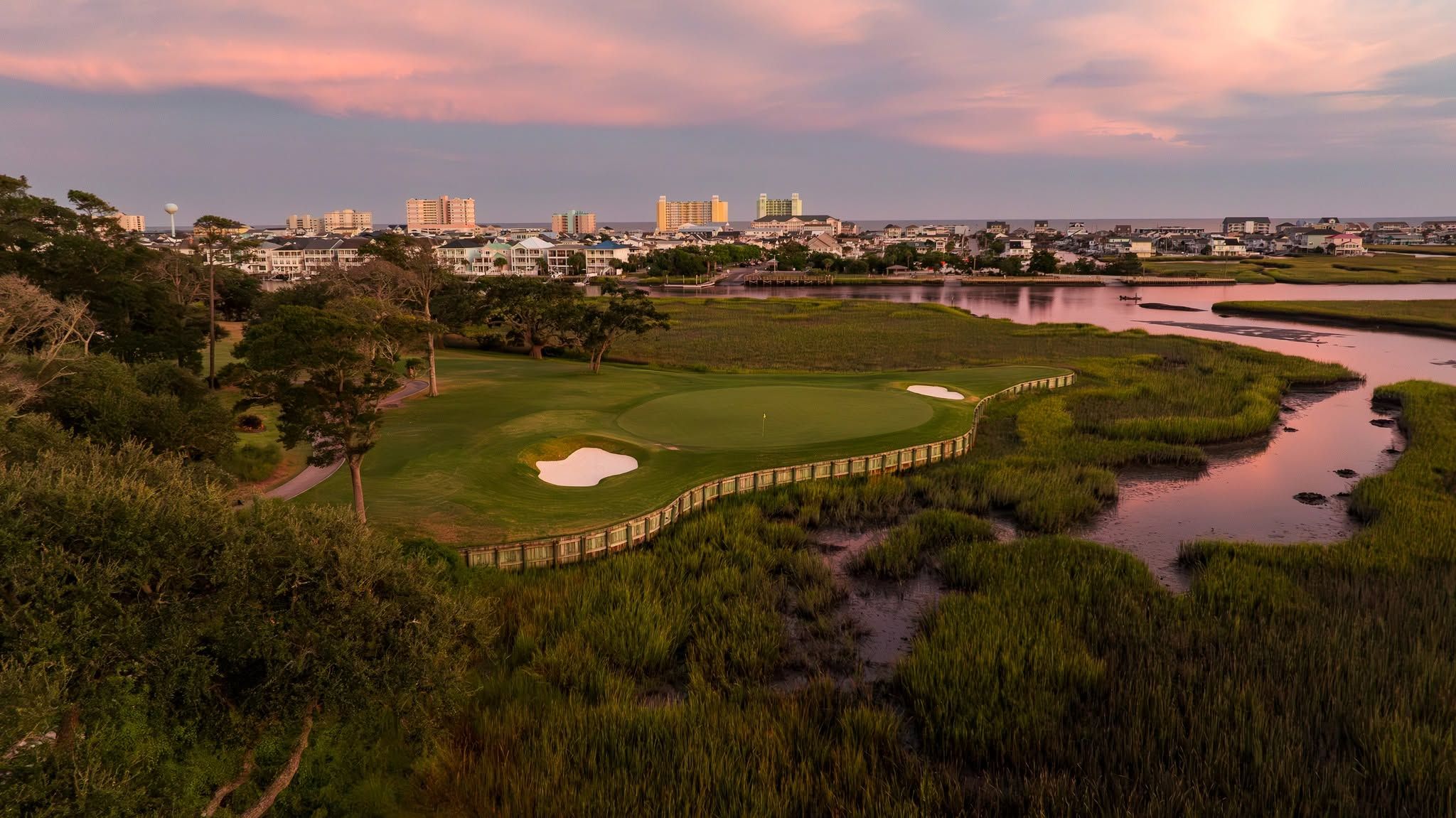 A sunset aerial of Tidewater Golf Club in North Myrtle Beach shows a green and bunker complex nestled between tidal marsh and a coastal waterway, with the oceanfront skyline glowing pink and purple in the distance.