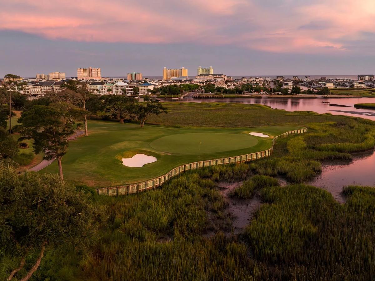 A sunset aerial of Tidewater Golf Club in North Myrtle Beach shows a green and bunker complex nestled between tidal marsh and a coastal waterway, with the oceanfront skyline glowing pink and purple in the distance.
