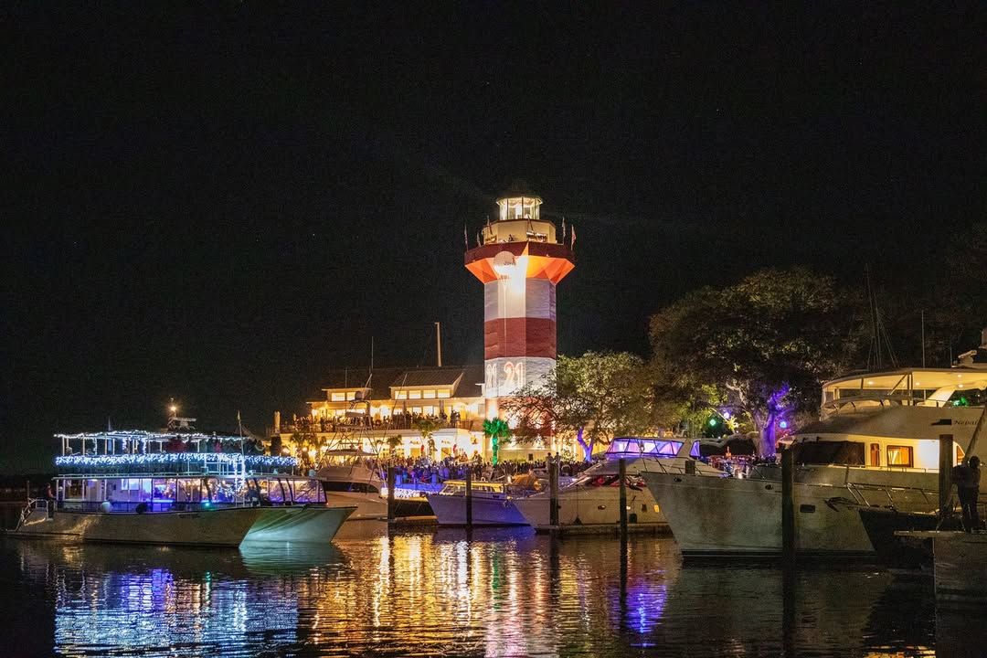 The iconic Harbour Town Lighthouse glows in red and white at night as boats fill the marina at Shelter Cove Towne Centre on Hilton Head Island during a New Year's Eve waterfront celebration, with festive lights reflecting across the water. 