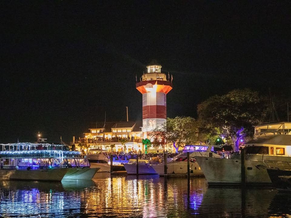 The iconic Harbour Town Lighthouse glows in red and white at night as boats fill the marina at Shelter Cove Towne Centre on Hilton Head Island during a New Year's Eve waterfront celebration, with festive lights reflecting across the water.