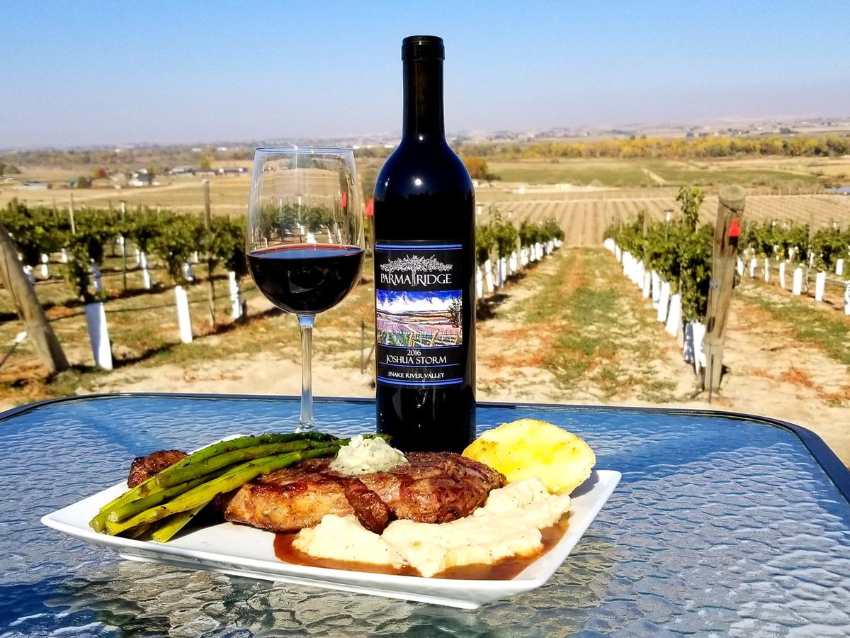 A bottle of Parma Ridge wine stands proudly on a glass table, paired with a hearty steak, mashed potatoes, and asparagus. Behind it, rows of vineyards stretch across the Snake River Valley under a clear sky. The setting blends fine dining with scenic vineyard views.