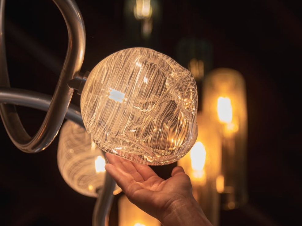 A hand gently holds a glowing glass light fixture with intricate white filament-like patterns, surrounded by other warm hanging bulbs.