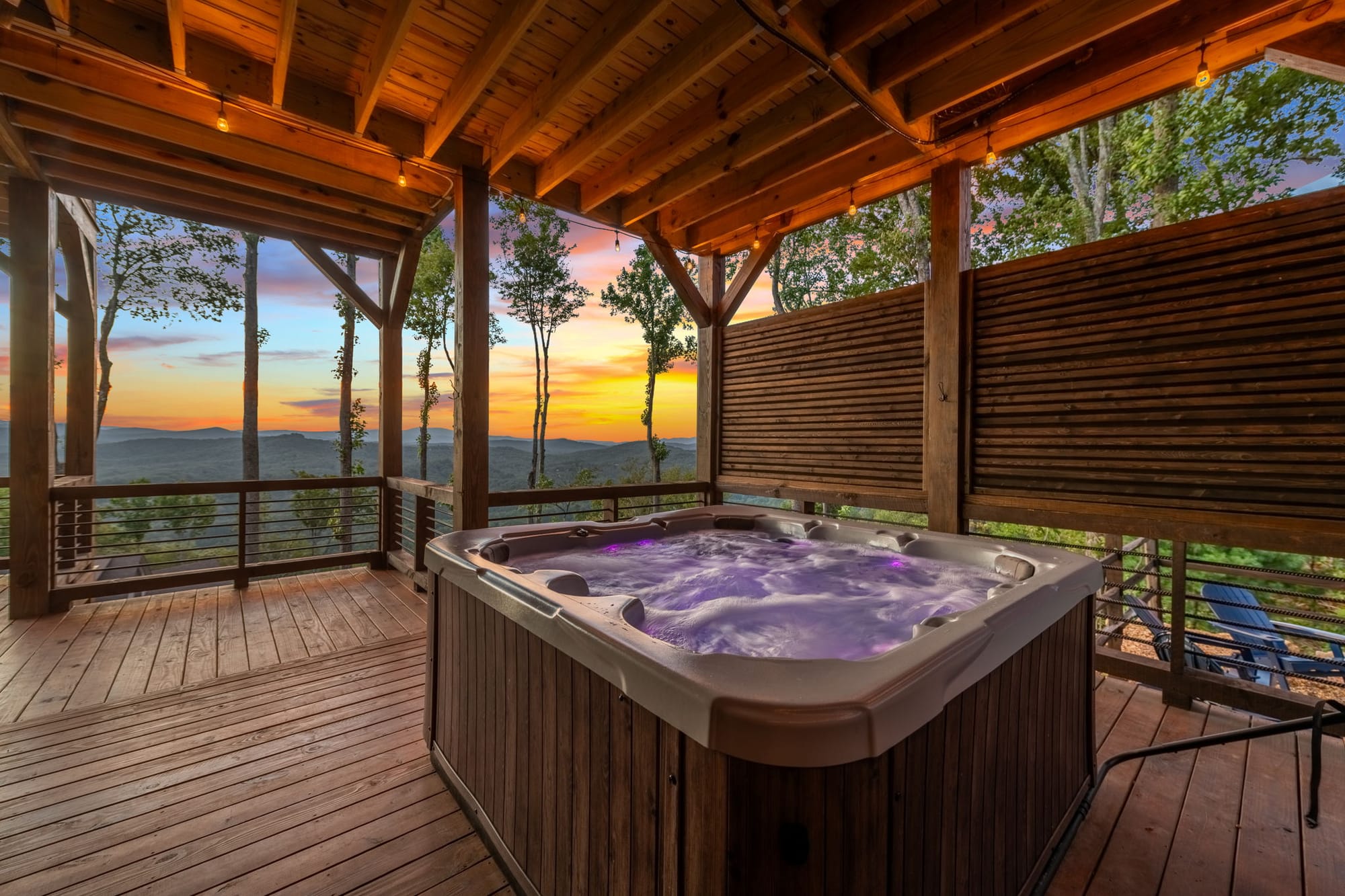 A bubbling hot tub sits on a wooden deck with string lights, facing a colorful mountain sunset. The view and warm water invite you to slow down, breathe, and reset your energy.