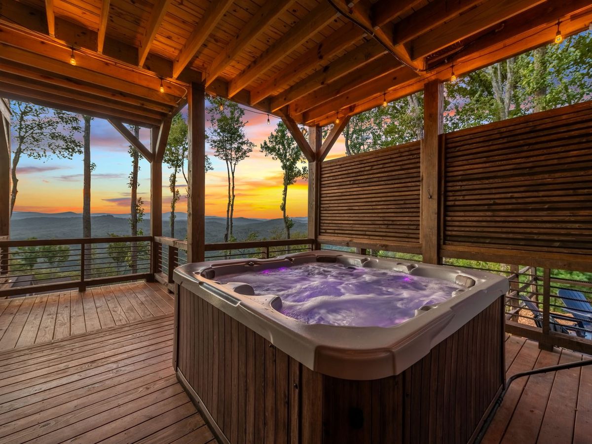 A bubbling hot tub sits on a wooden deck with string lights, facing a colorful mountain sunset. The view and warm water invite you to slow down, breathe, and reset your energy.