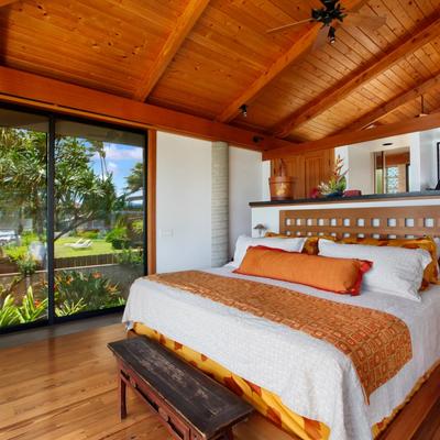 Bright, tropical-style bedroom with a large bed featuring orange and white bedding. The room has a high wooden ceiling and a glass wall offering lush garden and ocean views.