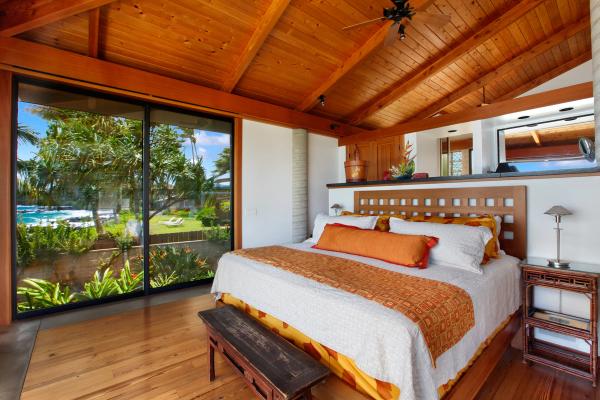 Bright, tropical-style bedroom with a large bed featuring orange and white bedding. The room has a high wooden ceiling and a glass wall offering lush garden and ocean views.