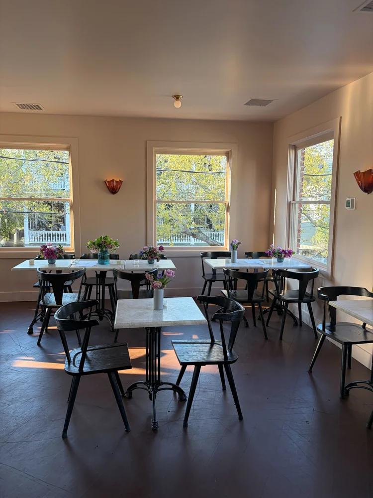 The bright, airy dining room at Baba's in Charleston, South Carolina, features white marble bistro tables with fresh floral arrangements, black bentwood chairs, warm amber sconce lighting, and large windows overlooking a leafy neighborhood street.