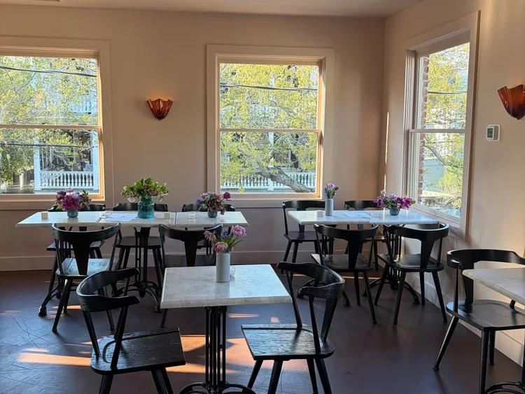 The bright, airy dining room at Baba's in Charleston, South Carolina, features white marble bistro tables with fresh floral arrangements, black bentwood chairs, warm amber sconce lighting, and large windows overlooking a leafy neighborhood street.