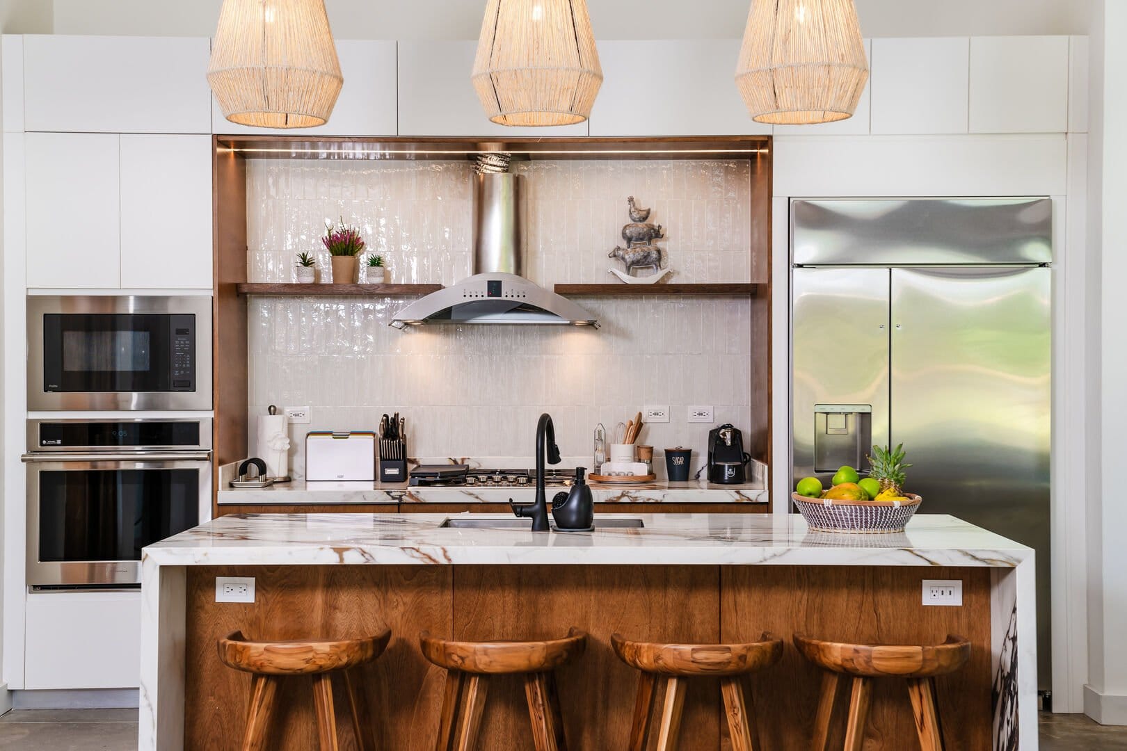 Modern kitchen with a marble island and wooden bar stools, stainless steel appliances, and woven pendant lights above.