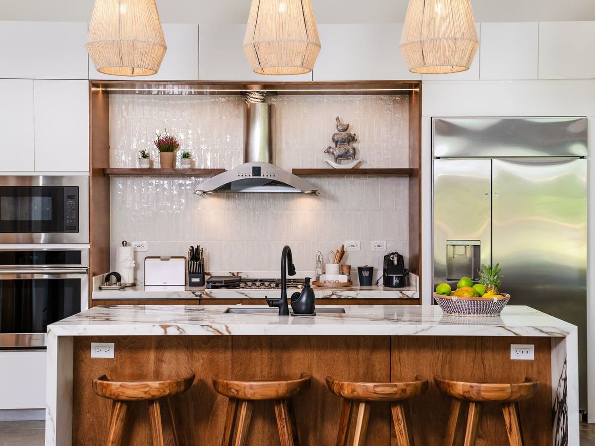 Modern kitchen with a marble island and wooden bar stools, stainless steel appliances, and woven pendant lights above.