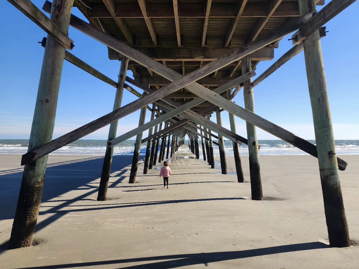 A child walks beneath a long wooden pier on a sunny beach, framed by crisscrossed beams and stretching shadows across the sand.