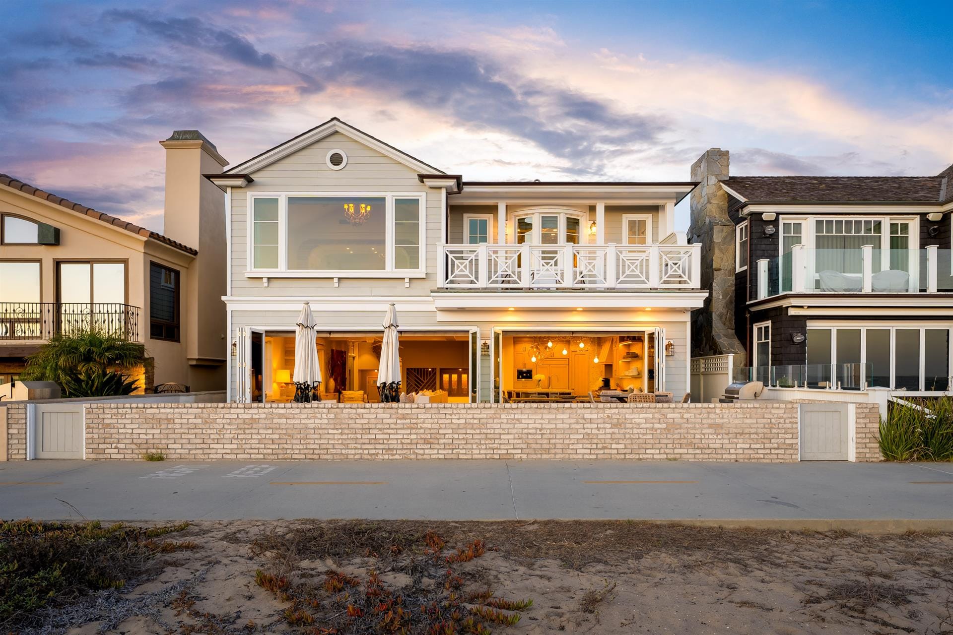 A beachfront vacation rental in Newport Beach, California, lit up at dusk with warm interior lights spilling through wide-open bi-fold garage doors facing the sand. The two-story coastal home features a second-floor balcony with white railings, a chandelier visible through the upper windows, and a relaxed California beach house feel.
