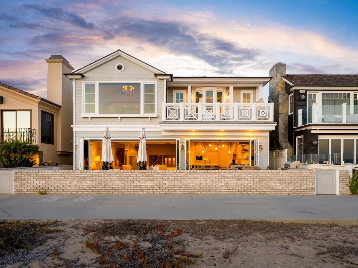 A beachfront vacation rental in Newport Beach, California, lit up at dusk with warm interior lights spilling through wide-open bi-fold garage doors facing the sand. The two-story coastal home features a second-floor balcony with white railings, a chandelier visible through the upper windows, and a relaxed California beach house feel.