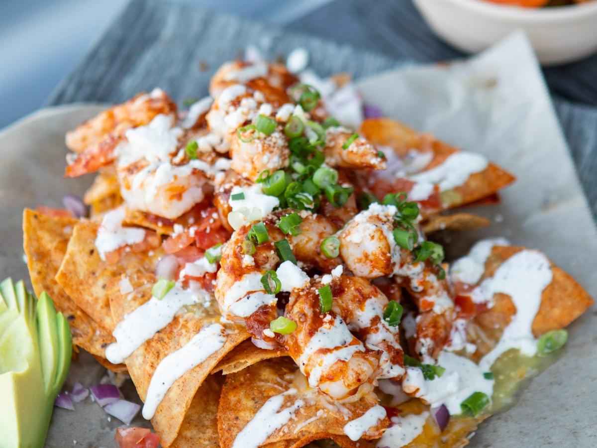 Close-up of crispy tortilla chips topped with spicy shrimp, melted cheese, fresh pico de gallo, crumbled cheese, and a drizzle of creamy sauce, garnished with chopped green onions and a side of avocado slices.
