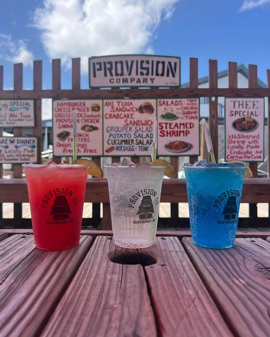 Red, white, and blue frozen drinks sit on a wooden table at The Provision Company with a seafood menu behind them. This casual waterfront spot is known for its fun drinks and laid-back vibe.