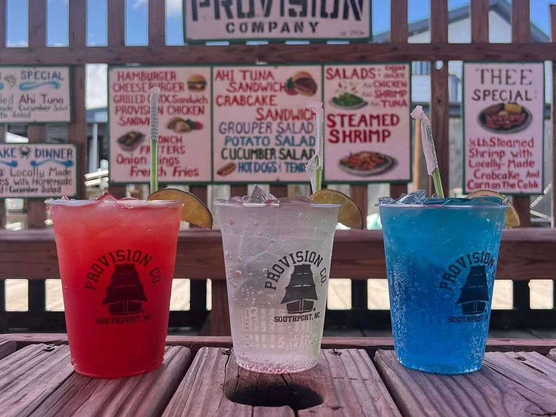 Red, white, and blue frozen drinks sit on a wooden table at The Provision Company with a seafood menu behind them. This casual waterfront spot is known for its fun drinks and laid-back vibe.