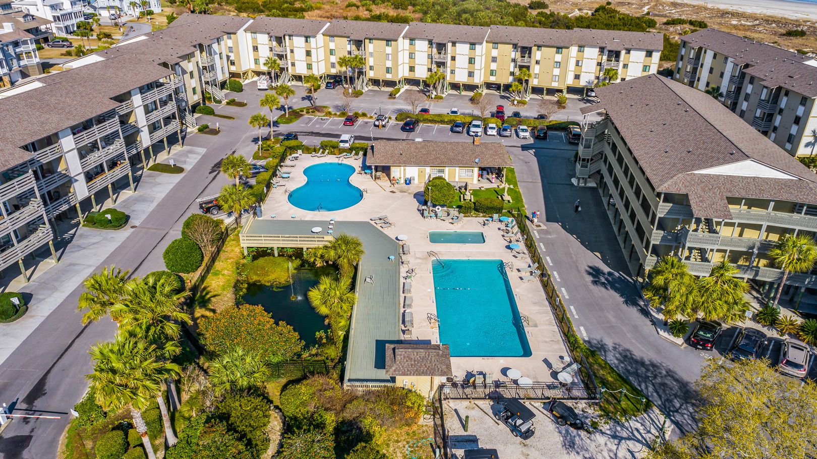 Aerial view of a beachfront condo complex featuring two outdoor swimming pools, a hot tub, lounge areas, and a landscaped pond, all within walking distance to the sandy shore.