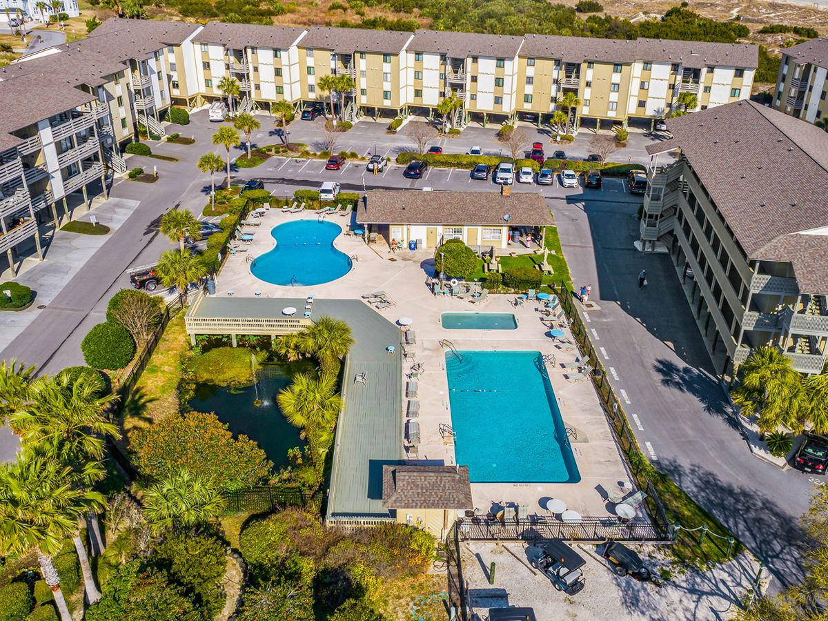 Aerial view of a beachfront condo complex featuring two outdoor swimming pools, a hot tub, lounge areas, and a landscaped pond, all within walking distance to the sandy shore.