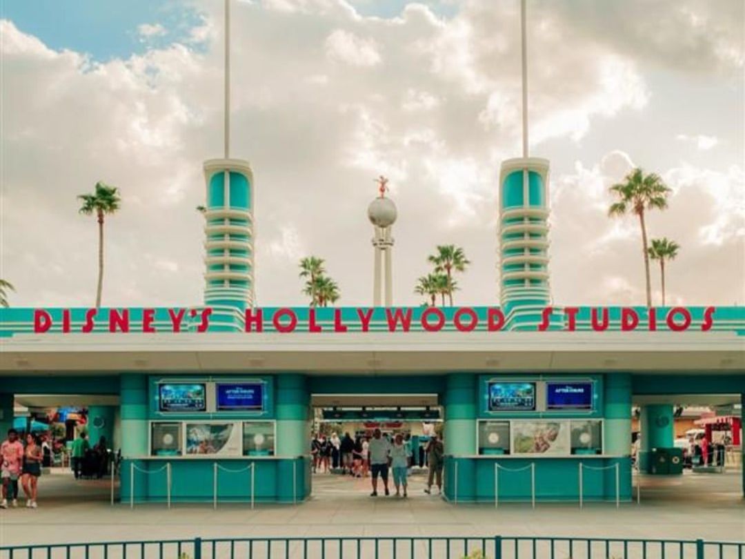 Entrance to Disney’s Hollywood Studios with palm trees, red flags, and retro-style towers under a partly cloudy sky. Visitors line up below the famous sign, ready for a day of shows, rides, and Disney magic.