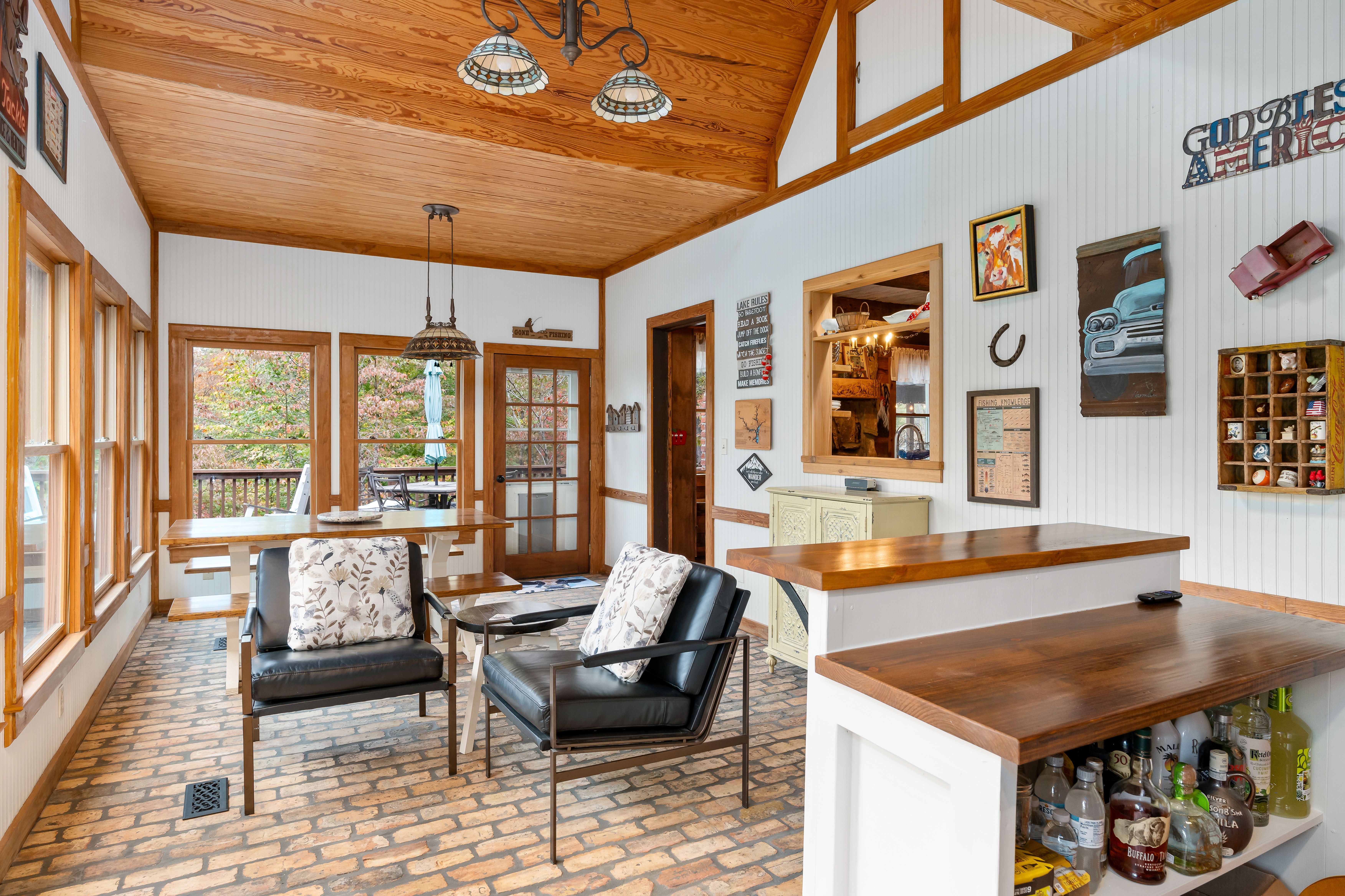 A rustic sunroom with brick flooring, wood-paneled ceiling, large windows, a wooden dining table, black leather chairs with floral pillows, and a small bar area with bottles displayed.