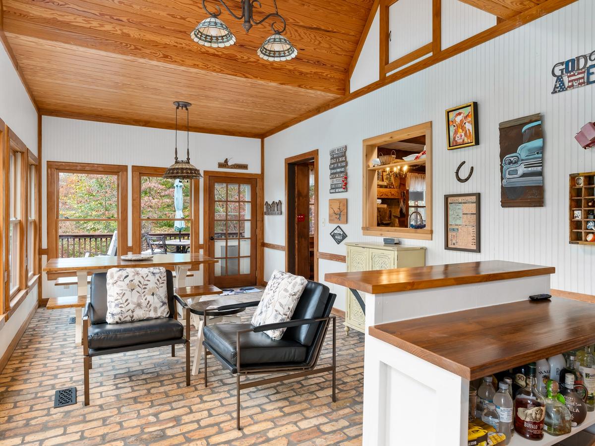 A rustic sunroom with brick flooring, wood-paneled ceiling, large windows, a wooden dining table, black leather chairs with floral pillows, and a small bar area with bottles displayed.